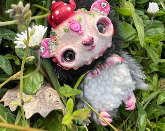 May include: A whimsical, handmade art doll of a possum-like creature with a red mushroom hat and floral accents. The doll has large, expressive eyes, a pink nose, and a fluffy white and gray body. It is nestled in green grass with white flowers.