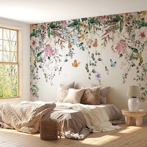 May include: A bedroom with a floral wallpaper featuring pink, yellow, and green flowers. The bed is made with a beige blanket and pillows. A small wooden table with a lamp sits next to the bed.