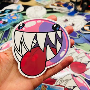 May include: A round sticker featuring a cartoon monster face with a wide grin, sharp white teeth, and a red tongue. The monster's face is a gradient of pink and purple, with black eyes and a white outline. Other stickers are in the background.