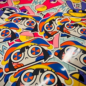 Op de afbeelding: Een verzameling kleurrijke stickers met cartoonfiguren met grote ogen, oranje en blauwe accenten en zwart haar. Andere stickers hebben roze, gele en paarse ontwerpen. De stickers liggen op een lichtblauwe ondergrond.