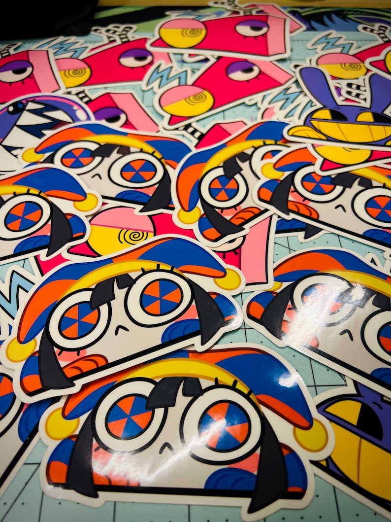 Op de afbeelding: Een verzameling kleurrijke stickers met cartoonfiguren met grote ogen, oranje en blauwe accenten en zwart haar. Andere stickers hebben roze, gele en paarse ontwerpen. De stickers liggen op een lichtblauwe ondergrond.