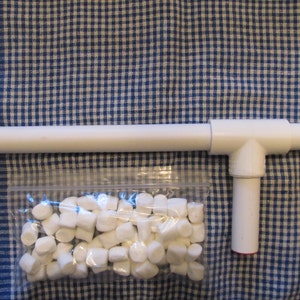 May include: A white PVC pipe structure with a T-shaped connector and a small bag of white marshmallows. The structure has a red cap on the bottom. The items are on a blue and white checkered fabric.