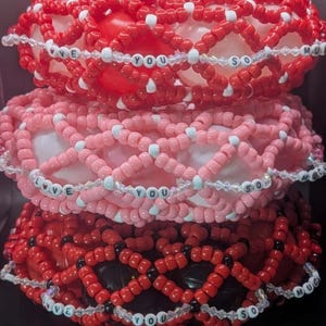 May include: Three-tiered beaded art piece in shades of red and pink. Each tier features a lattice design with the words "LOVE YOU SO MUCH" spelled out in white beads. The piece is a decorative item.