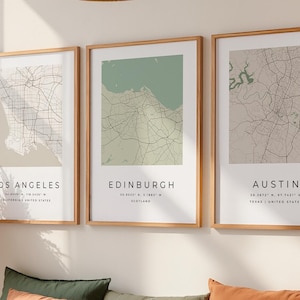 May include: Three framed city map prints hang above a bed with decorative pillows and a rust-colored throw. The prints feature maps of Los Angeles, Edinburgh, and Austin, with city names and coordinates.
