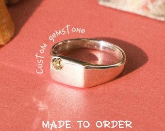 May include: A silver signet ring featuring a single small round yellow gemstone set into the flat rectangular face. The text "Custom gemstone" and "MADE TO ORDER" is printed in white near the ring on a coral-colored surface.