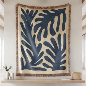 May include: A large, woven throw blanket with a cream background and navy blue abstract leaf design. The blanket has a fringed edge with red, brown, and cream accents. The throw is displayed on a wall, adding a decorative touch to the room.