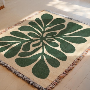 May include: A woven throw blanket with a large, stylized green floral design on a cream background. The blanket has a fringed edge with a mix of colors. The design is symmetrical and modern, suitable for home decor.
