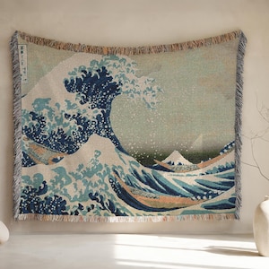 May include: Woven tapestry throw blanket featuring the iconic "Great Wave off Kanagawa" artwork. The design showcases a large wave in shades of blue and white, with boats and a mountain in the background. The blanket has fringed edges and is displayed on a wall.