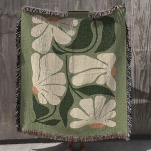 May include: A green woven throw blanket featuring a floral design with large white flowers and dark green leaves. The flowers have peach-colored centers. The blanket has a fringed edge in shades of brown, tan, and black.