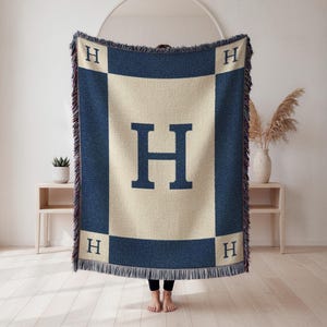 May include: A navy blue and beige throw blanket with the letter "H" prominently displayed. The blanket features a large "H" in the center and smaller "H" letters in each corner. The edges are fringed.