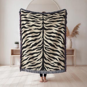 May include: A rectangular woven throw blanket featuring a black and cream tiger stripe pattern. The blanket has a textured surface and is finished with decorative fringe along the top and bottom edges, creating a distinct animal-inspired design.
