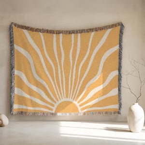 May include: A woven throw blanket with a yellow and white sunburst design. The blanket features a fringe border and is displayed against a neutral wall. The sunburst pattern radiates from the bottom center.