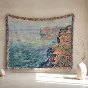 May include: A woven tapestry wall hanging featuring a landscape scene of a rocky cliff overlooking a body of water. The water is a light blue and green color, and the sky is a light blue. The cliff is a brown and orange color, and there are some trees on the cliff.