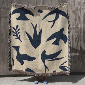 May include: A woven throw blanket in a light beige color featuring a repeating pattern of dark navy blue swallows and leafy branch silhouettes. The rectangular textile has a textured surface and is finished with decorative fringed edges.