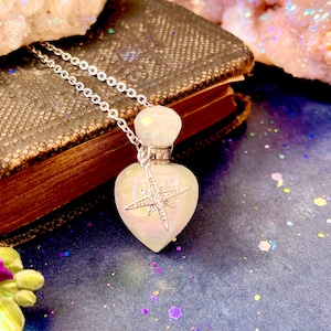 May include: A white iridescent heart-shaped bottle with a silver star charm on a silver chain necklace. The bottle is on a dark blue surface with iridescent glitter.