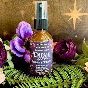 May include: A clear glass spray bottle with a black nozzle and a purple label featuring the text Anita Apothecary, Botanical Magick, Empath, Ground & Protect, Hydrosol Mist with Black Tourmaline & Amethyst Crystals Infused with Essential Oils of Lavender & Rosemary.