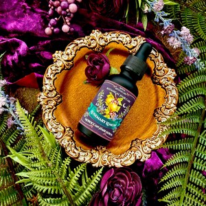 Pode incluir: Uma garrafa preta de s&eacute;rum capilar com conta-gotas, rotulada como "Rosemary Lemon Moroccan Hair Serum", est&aacute; sobre um prato dourado e ornamentado. O prato repousa sobre veludo roxo, rodeado por flores roxas escuras e fetos verdes. Est&eacute;tica luxuosa e bot&acirc;nica.