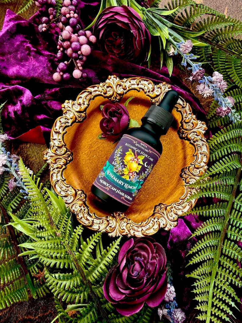 Pode incluir: Uma garrafa preta de s&eacute;rum capilar com conta-gotas, rotulada como "Rosemary Lemon Moroccan Hair Serum", est&aacute; sobre um prato dourado e ornamentado. O prato repousa sobre veludo roxo, rodeado por flores roxas escuras e fetos verdes. Est&eacute;tica luxuosa e bot&acirc;nica.