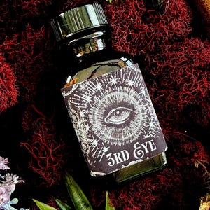 May include: A dark glass bottle with a black cap and a label that reads "3RD EYE". The label features a detailed eye illustration surrounded by radiating lines and stars. The bottle is set against a backdrop of red moss and greenery.