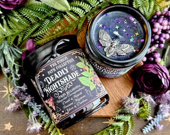 May include: Two black glass jar candles from The Poison Path Collection. One candle is labeled "Deadly Nightshade" with text and botanical illustrations. The other candle has a silver moth embellishment. Both candles are surrounded by purple flowers and greenery.