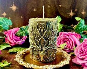 May include: A gold candle with a detailed design of a stag head and intricate floral patterns. The candle is sitting on a gold metal tray.