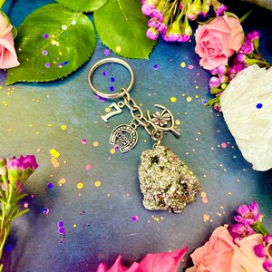 Pode incluir: Um chaveiro de prata com uma ferradura, um trevo de quatro folhas e um pequeno amuleto de pedra cinza brilhante. O chaveiro est&aacute; rodeado de flores rosa e roxas.