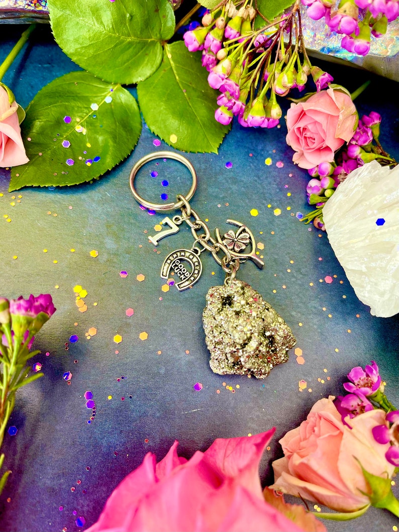 Pode incluir: Um chaveiro de prata com uma ferradura, um trevo de quatro folhas e um pequeno amuleto de pedra cinza brilhante. O chaveiro est&aacute; rodeado de flores rosa e roxas.