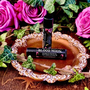 May include: Two black lip balm tubes with gold lettering on a gold and white decorative plate. The lip balm tubes are labeled "Botanical Beauty Blood Moon Quartz Crystal Infused Lip Balm". The plate is surrounded by green ivy leaves and pink roses.