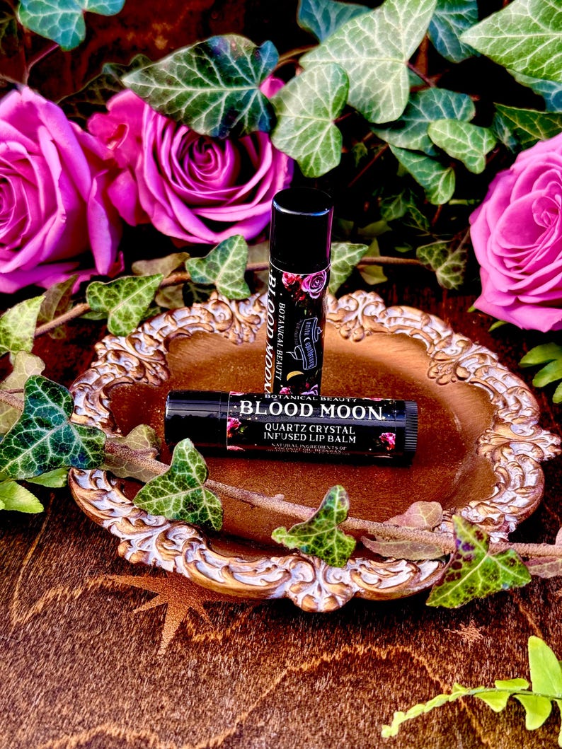 May include: Two black lip balm tubes with gold lettering on a gold and white decorative plate. The lip balm tubes are labeled "Botanical Beauty Blood Moon Quartz Crystal Infused Lip Balm". The plate is surrounded by green ivy leaves and pink roses.
