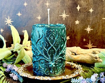 May include: A teal-colored, cylindrical candle with an embossed deer design. The candle sits on a decorative gold-colored plate, surrounded by greenery and small purple flowers. The background features a starry pattern.