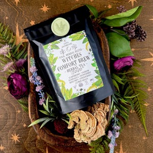 May include: A black bag of "Witches Comfort Brew - Herbal Tea" with a green seal, surrounded by greenery and flowers. The tea blend includes elderberry, yarrow, echinacea, peppermint, rose hips, ginger root, and lemongrass. The packaging is set on a wooden surface.