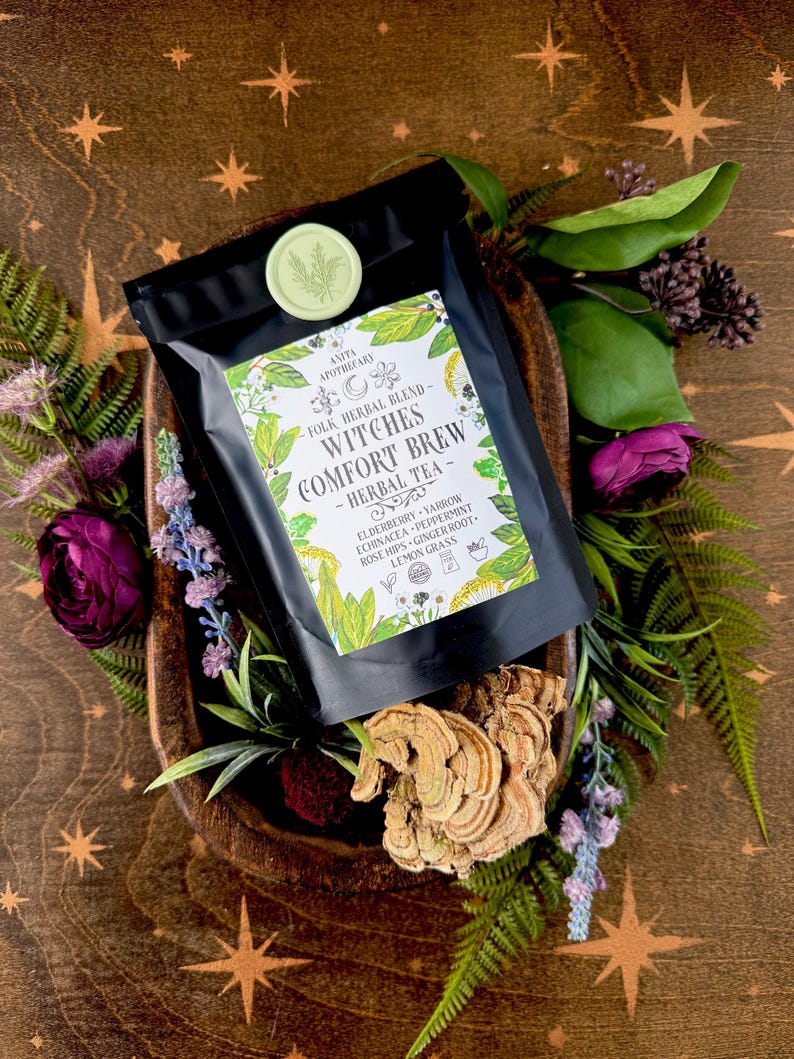 May include: A black bag of "Witches Comfort Brew - Herbal Tea" with a green seal, surrounded by greenery and flowers. The tea blend includes elderberry, yarrow, echinacea, peppermint, rose hips, ginger root, and lemongrass. The packaging is set on a wooden surface.