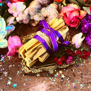 May include: A bundle of Palo Santo sticks tied with twine and a purple ribbon, resting on a gold ornate tray. The sticks are surrounded by pink and purple flowers, a crystal, and colorful confetti.