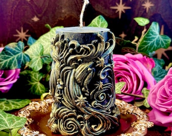 May include: A black and gold candle with a detailed, ornate design. The candle is decorated with swirling patterns and stars. It is sitting on a gold-colored decorative plate.