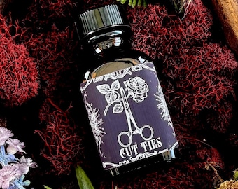 May include: A dark glass bottle with a black cap and a label that reads "CUT TIES." The label features a rose, scissors, and floral designs in white against a dark purple background. The bottle is surrounded by red moss and greenery.