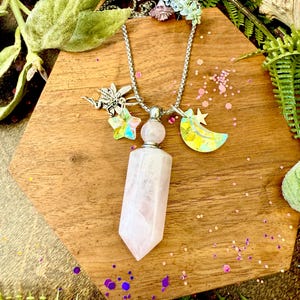 May include: A silver chain necklace with a pink crystal pendant and iridescent star and moon charms. The necklace is displayed on a wooden surface with floral and leaf accents, creating a whimsical, bohemian aesthetic.