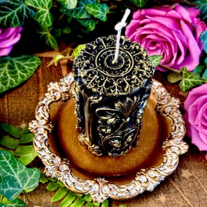 May include: A black and gold decorative pillar candle with intricate floral designs. The candle is sitting on a gold and white decorative plate.