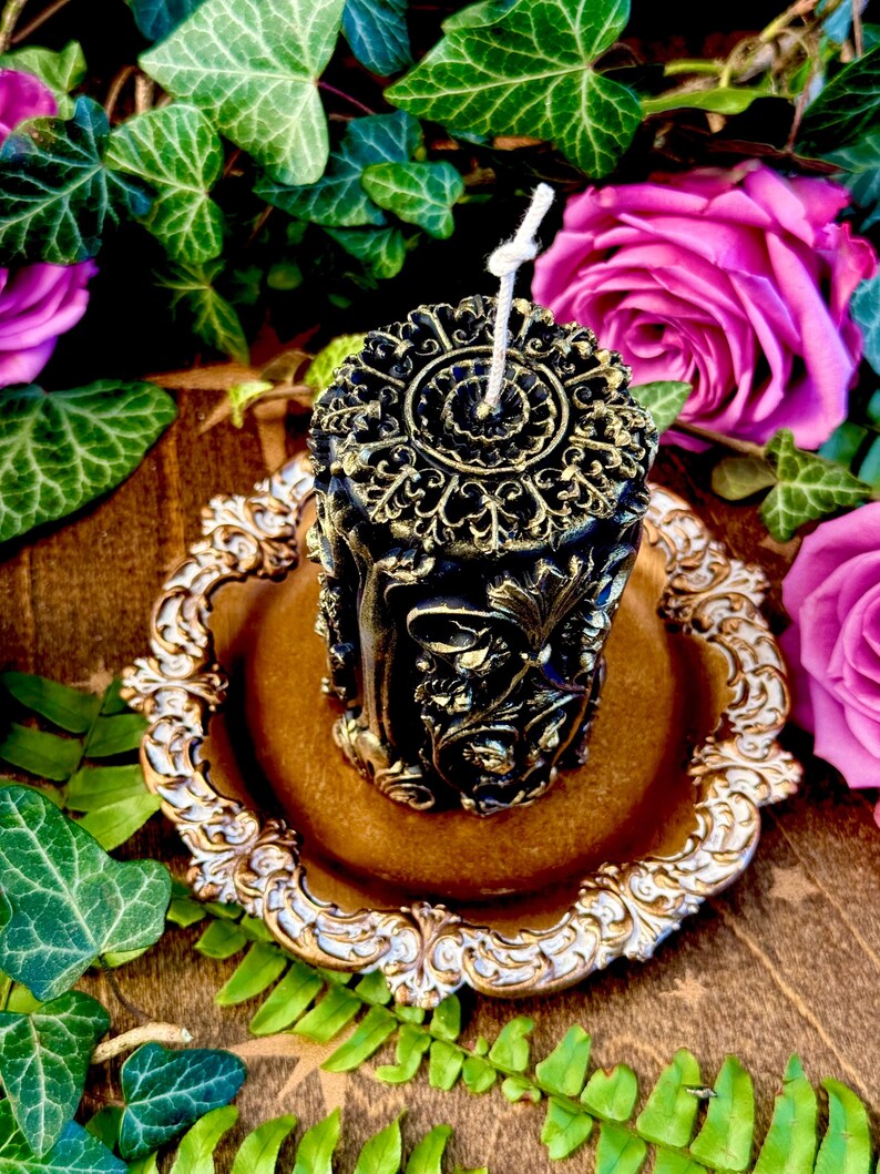 May include: A black and gold decorative pillar candle with intricate floral designs. The candle is sitting on a gold and white decorative plate.