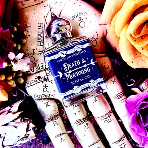 Pu&ograve; includere: Un piccolo flacone di vetro blu con un tappo argentato e un'etichetta che recita "Anita Apothecary Death & Mourning Ritual Oil". Il flacone &egrave; appoggiato su una mano con simboli astrologici e circondato da fiori.