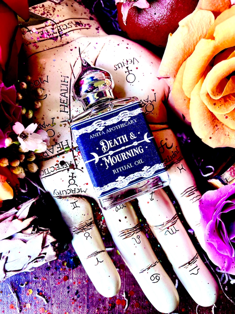 Pu&ograve; includere: Un piccolo flacone di vetro blu con un tappo argentato e un'etichetta che recita "Anita Apothecary Death & Mourning Ritual Oil". Il flacone &egrave; appoggiato su una mano con simboli astrologici e circondato da fiori.