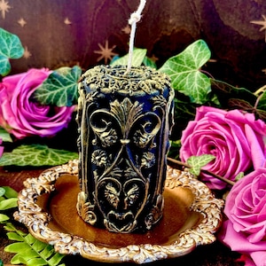 May include: A black and gold decorative candle with an ornate design. The candle is sitting on a gold-colored plate.