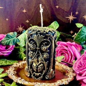 May include: A black and gold decorative pillar candle with an intricate design. The candle is sitting on a gold tray.