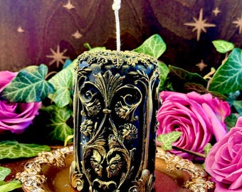 May include: A black and gold decorative pillar candle with an intricate design. The candle is sitting on a gold tray.