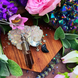 May include: A silver necklace with a pendant featuring a crescent moon, stars, and a black stone crescent moon. A brown crystal vial pendant hangs next to the moon pendant. The necklace rests on a wooden surface surrounded by flowers and crystals.