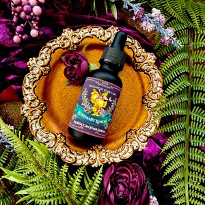 Pode incluir: Uma garrafa preta de "S&eacute;rum capilar marroquino de alecrim e lim&atilde;o" com conta-gotas, colocada em um prato dourado ornamentado. O fundo apresenta veludo roxo, flores e vegeta&ccedil;&atilde;o, criando um tema bot&acirc;nico. O r&oacute;tulo diz "Anita Apothecary Botanical Hair Care".