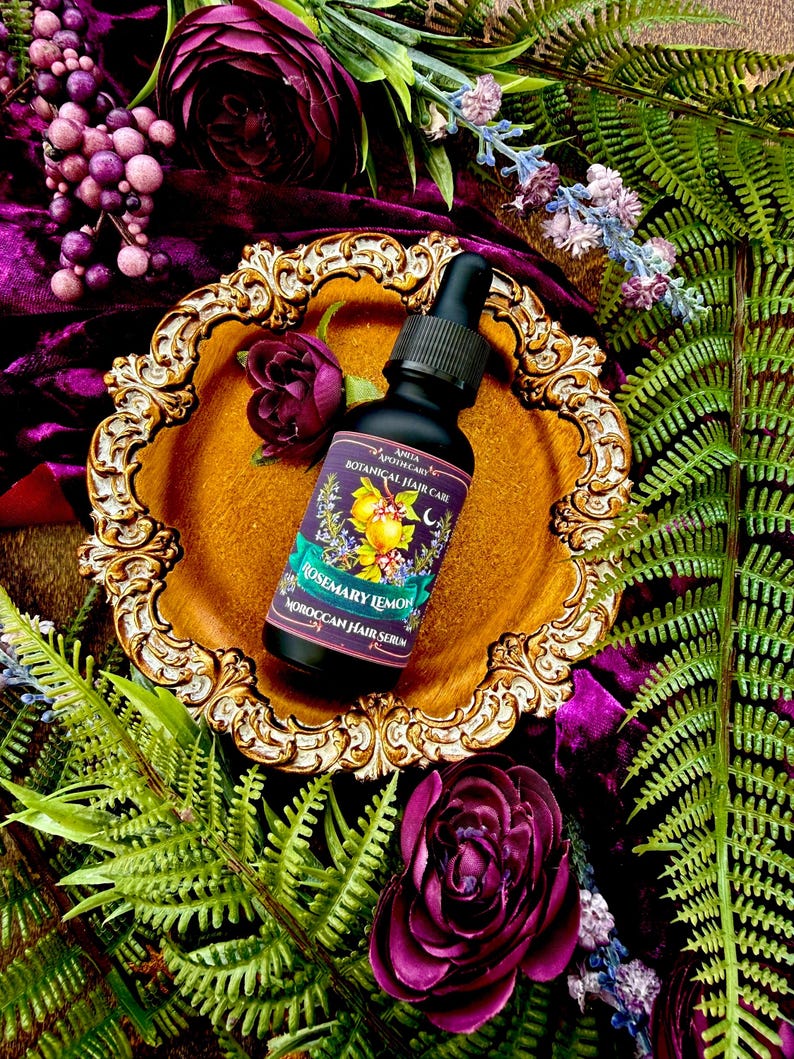 Pode incluir: Uma garrafa preta de "S&eacute;rum capilar marroquino de alecrim e lim&atilde;o" com conta-gotas, colocada em um prato dourado ornamentado. O fundo apresenta veludo roxo, flores e vegeta&ccedil;&atilde;o, criando um tema bot&acirc;nico. O r&oacute;tulo diz "Anita Apothecary Botanical Hair Care".
