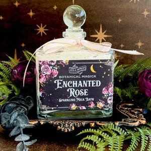 May include: A clear glass bottle with a glass stopper and a pink ribbon, filled with a sparkling milk bath. The label reads "Enchanted Rose" with floral and celestial designs. The bottle sits on a decorative stand.