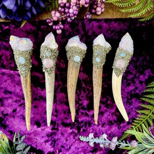 May include: Five decorative antler wands with crystal and gemstone embellishments. Each wand features a unique combination of crystals, pyrite, and gemstones in shades of white, pink, and blue. The wands are set against a vibrant purple velvet background.