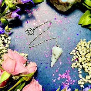 May include: A silver pendulum with a pentagram charm hangs from a chain on a dark blue background with pink and purple glitter.  The pendulum is made of a clear crystal with a pointed tip.  There are pink roses, purple flowers, and a white crystal cluster in the background.