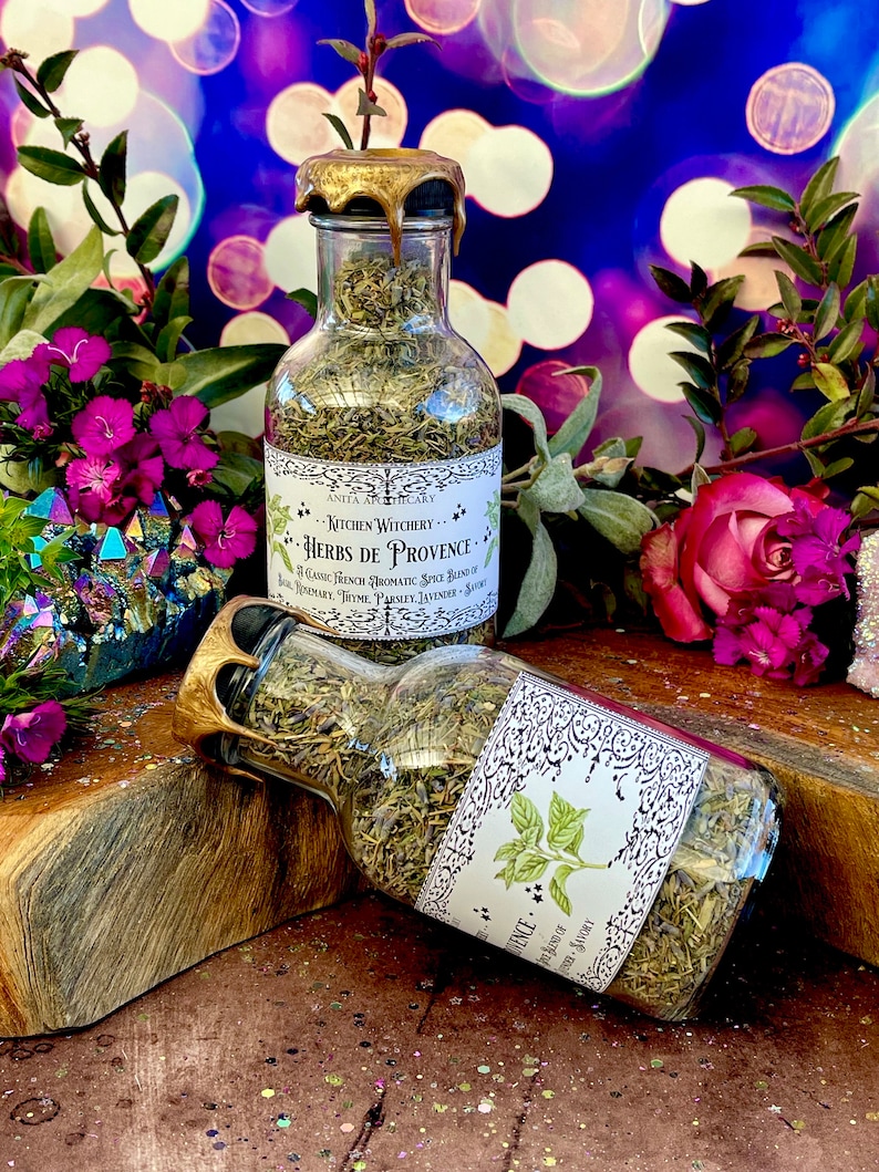May include: A glass bottle filled with dried herbs, labeled "Kitchen Witchery Herbs de Provence." The bottle has a gold lid and is sitting on a wooden surface.